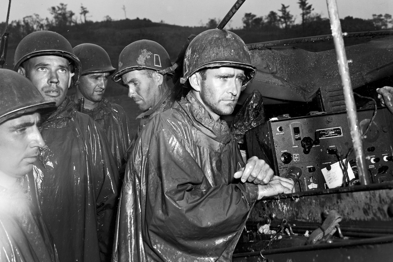 Five men wearing helmets and rain ponchos stand near a radio to hear a broadcast.