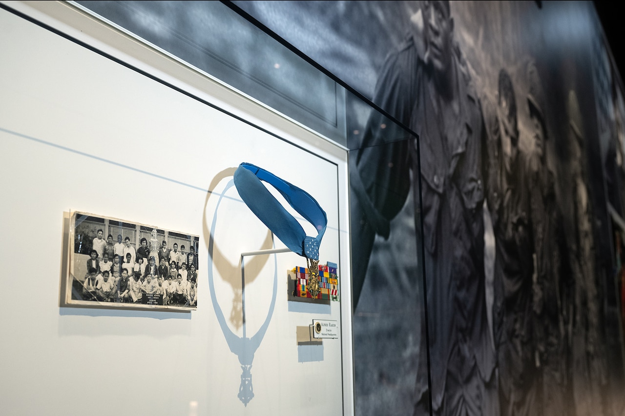A medal on a blue ribbon, a military ribbon rack and an old photo are displayed in a glass case.