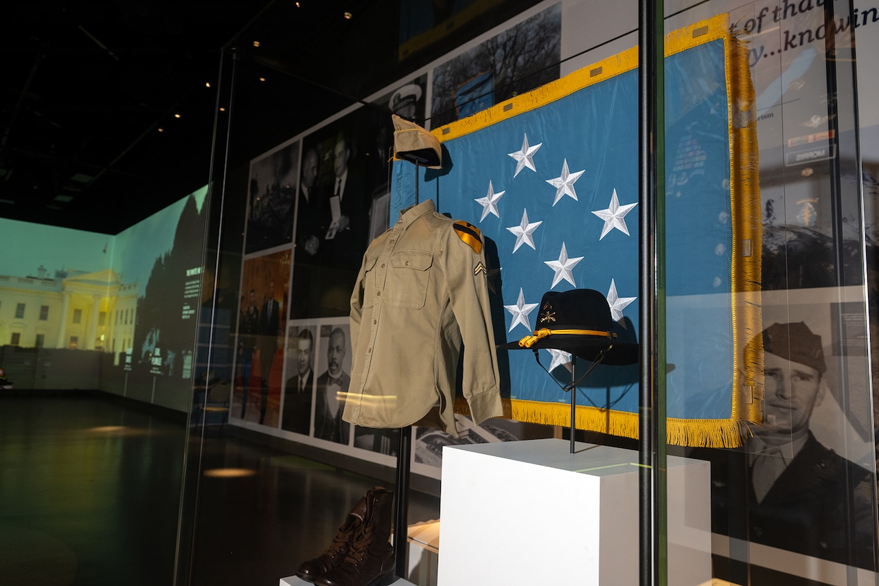 A blouse and a hat hang in front of an old flag in a display case.