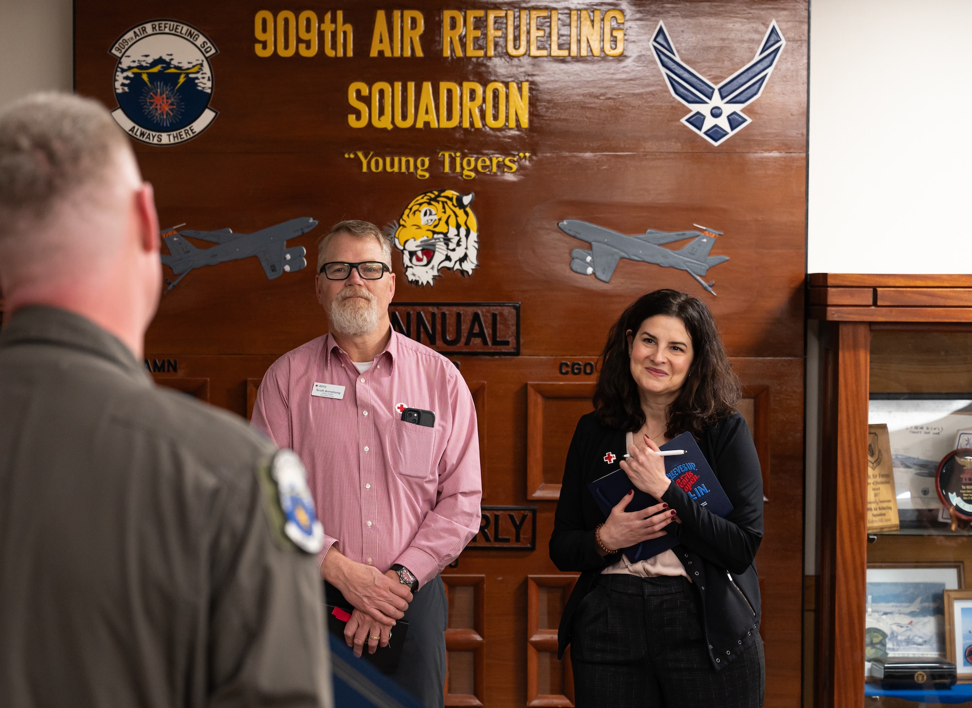 A man and a woman (both in focus) listen to a male Airman in uniform (slightly out of focus) give a briefing.