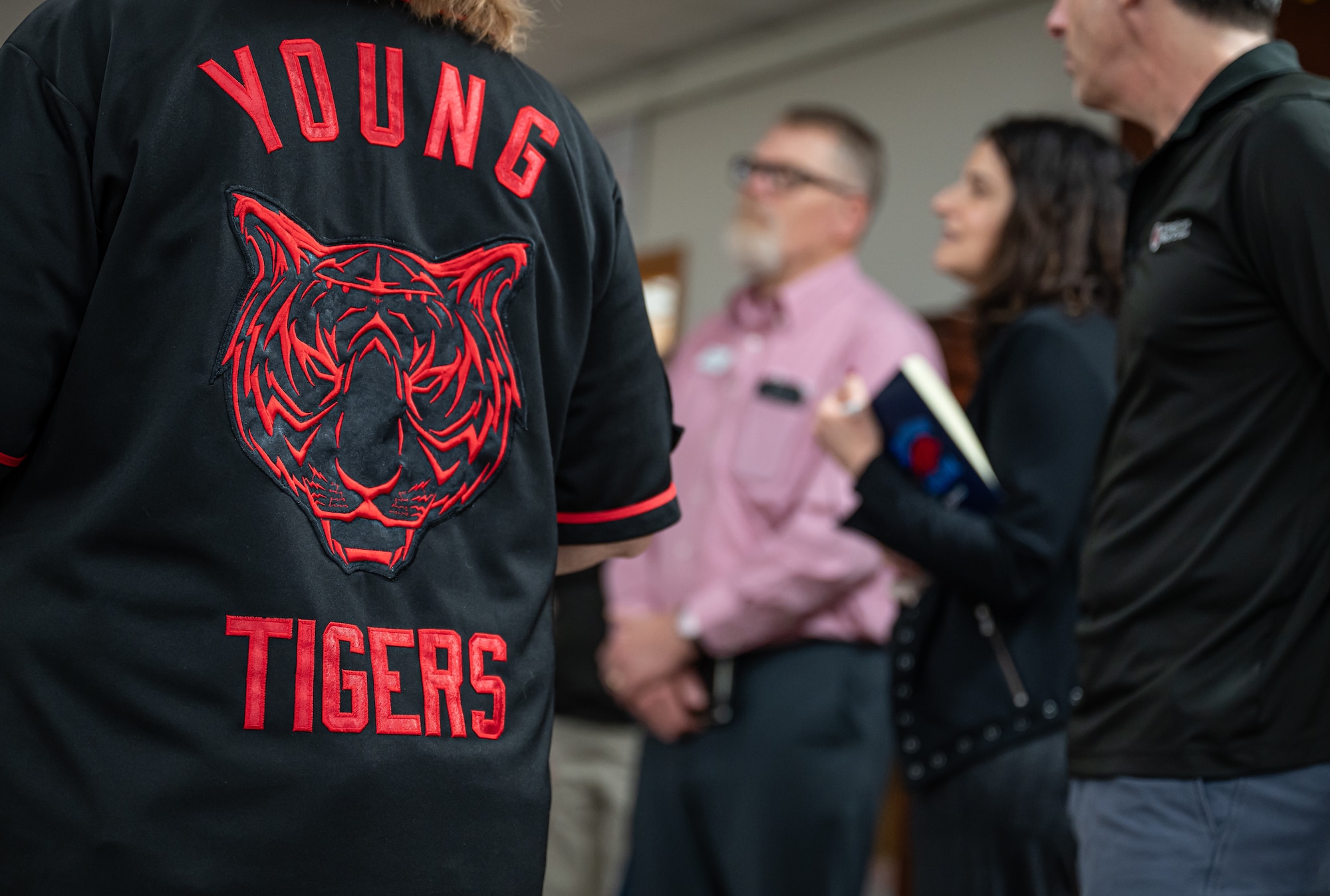 A black jersey with a red tiger head outline and the text "Young Tiger" is worn by a person in focus in the fore ground while three people in the background stand listening to a briefing out of focus.