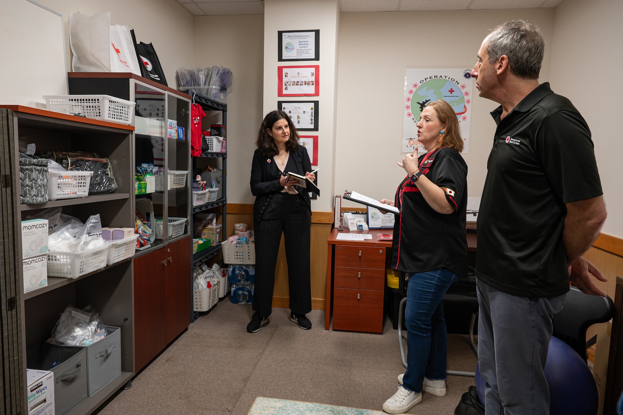 A woman holding a clip board, gives a briefing to two other people in the room. The room has several shelves neatly stocked with toys, nursing supplies and other items.