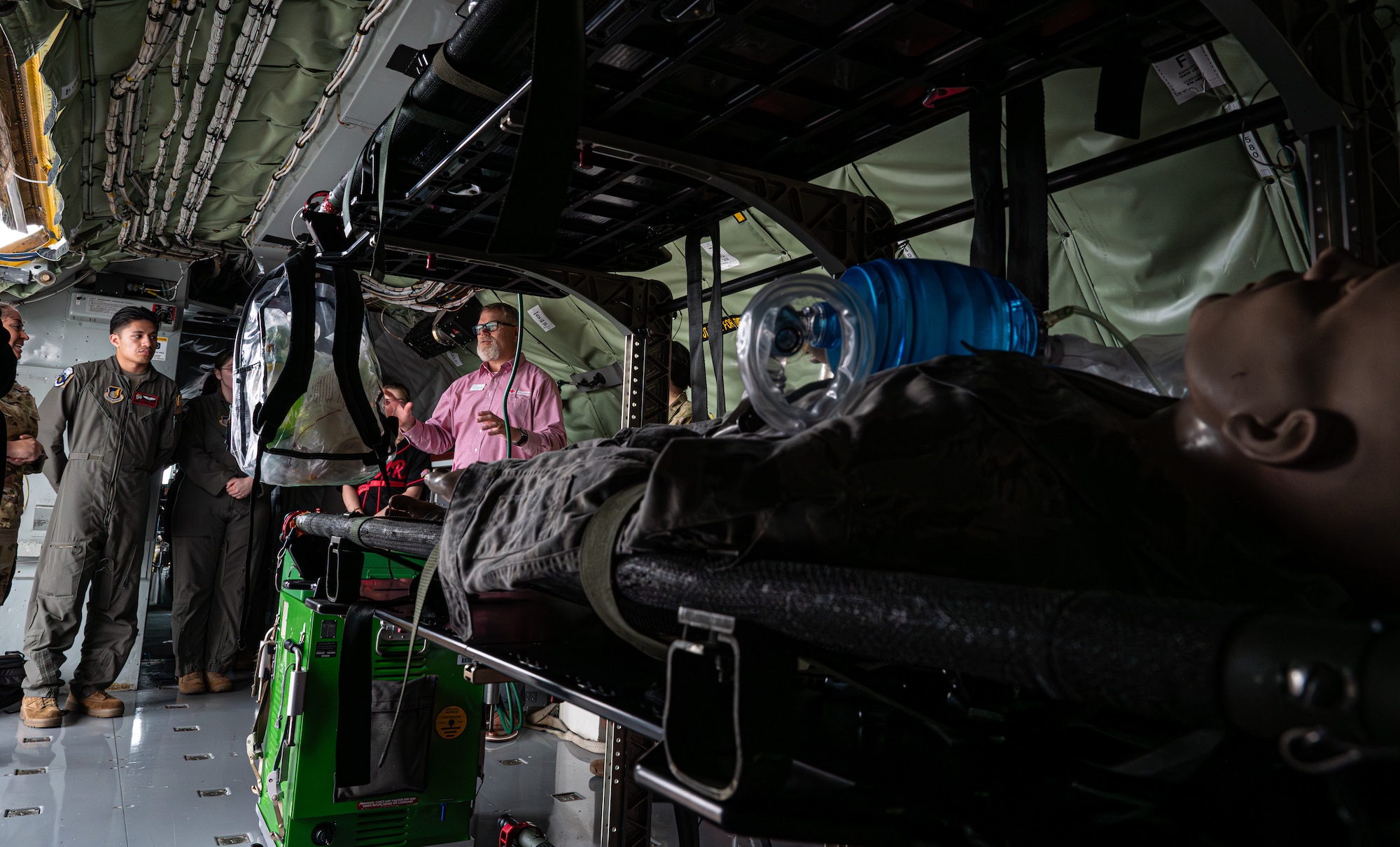 A man in a pink shirt talks to people inside a KC-135 Stratotanker. The photo is taken at a lower angle and in the foreground is a manakin strapped to a litter as a simulated patient.