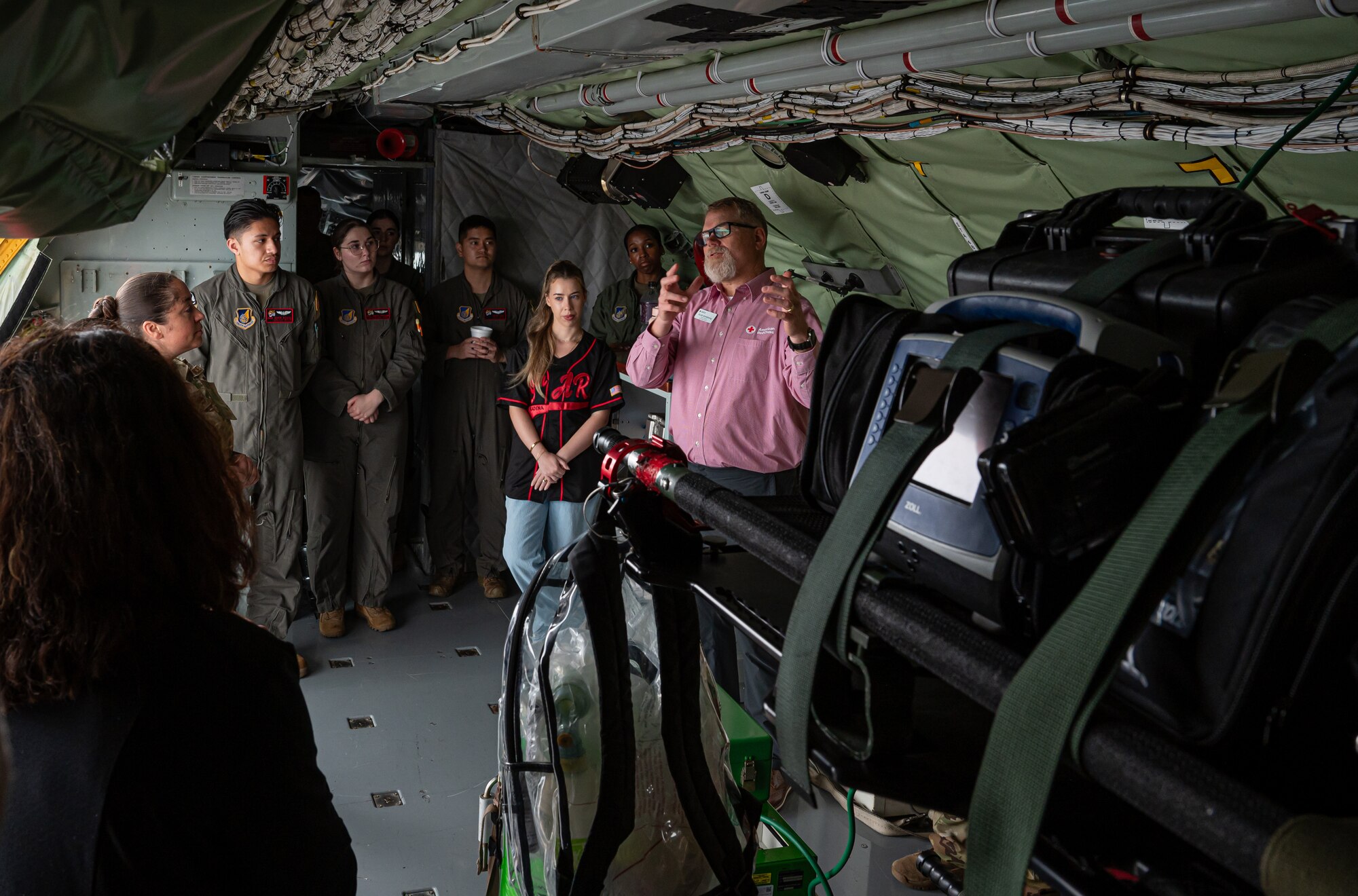A man in a pink shirt talks to a group of people in a KC-135 Stratotanker. In the foreground of the image lies medical equipment strapped to a litter.