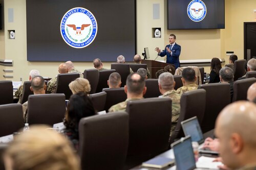 A man in a business suit is standing behind a podium gesturing with his hands. In front of him, roughly two dozen people are seated, watching him speak.