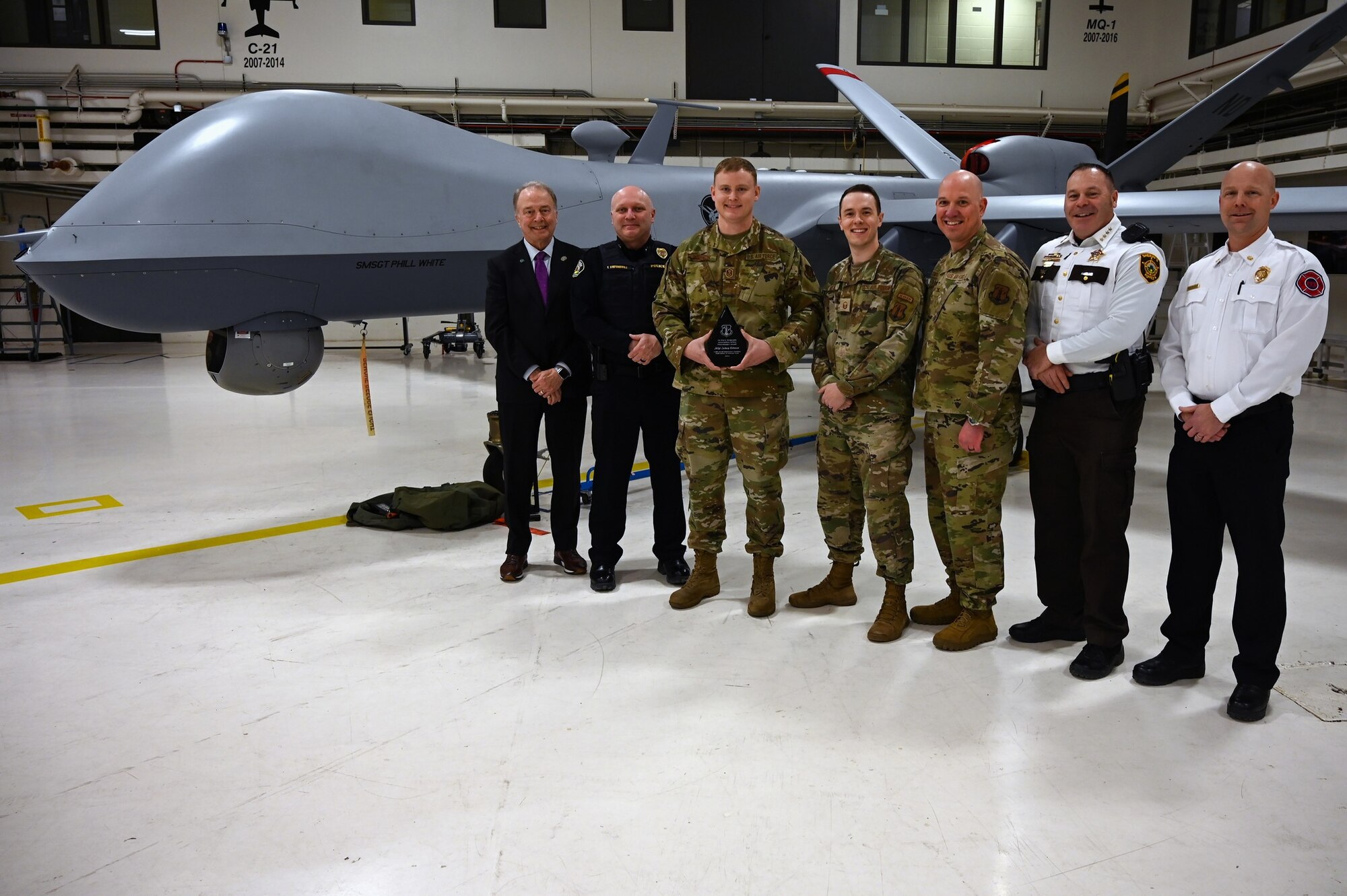 Uniformed service members and civilians pose for a group photo while the uniformed service member three people from the left holds up an award. The people stand in front of MQ-9 Reaper aircraft in a hangar.