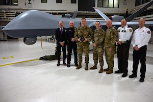 Uniformed service members and civilians pose for a group photo while the uniformed service member three people from the left holds up an award. The people stand in front of MQ-9 Reaper aircraft in a hangar.