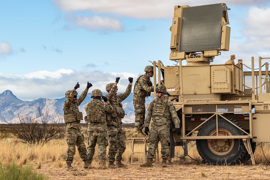 Four soldiers stand and signal next to radar equipment while another works on a laptop attached to the radar equipment.