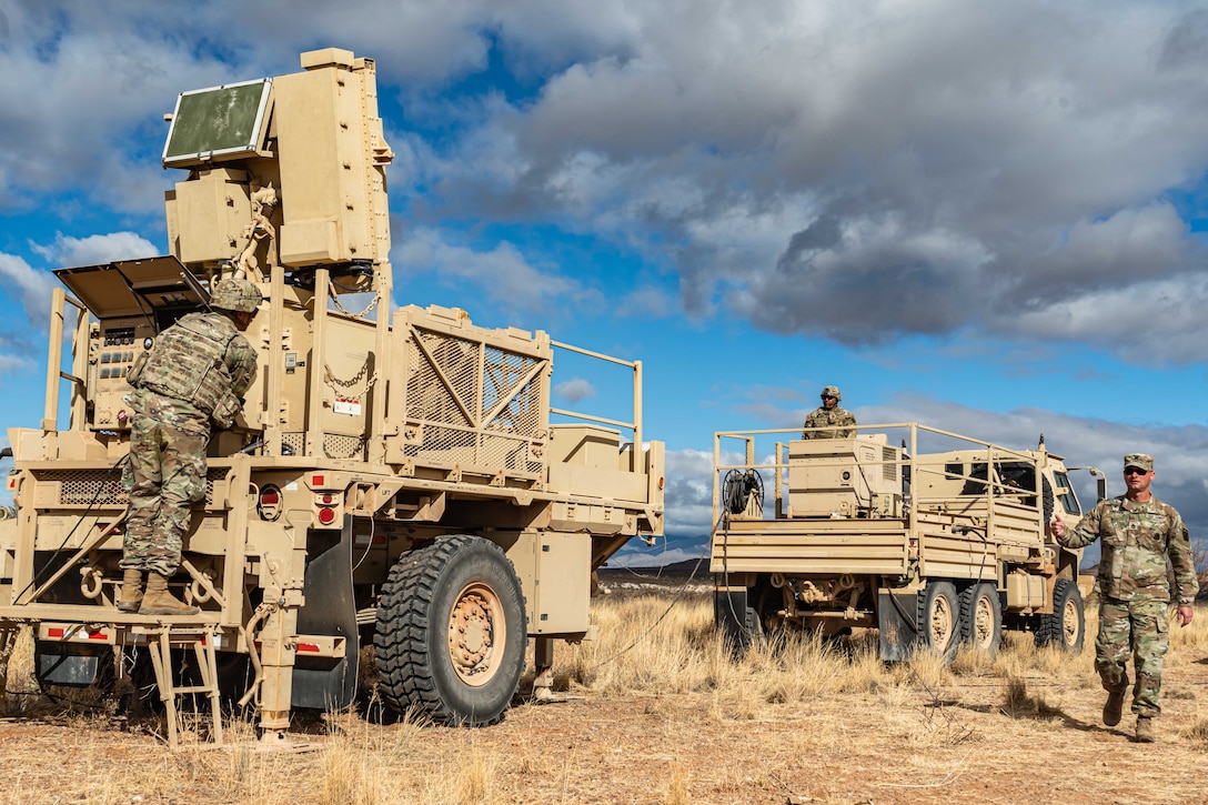 A soldier presides over radar system equipment while another soldier walks nearby on an arid field.