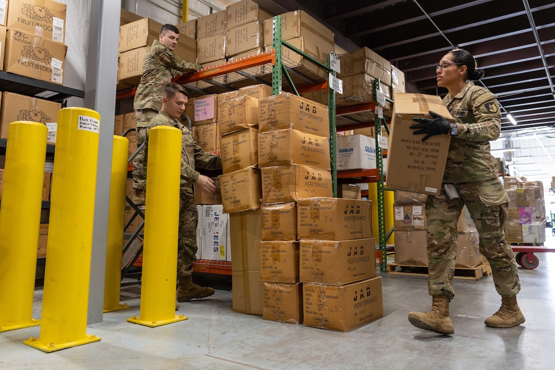 Three soldiers stack boxes inside a warehouse.
