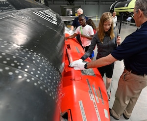 a girl and a boy, both holding a white canes and wearing white gloves run their hands down the side of a red and black fuselage while 2 museum volunteers wearing blue shirts and khaki pants speak to them