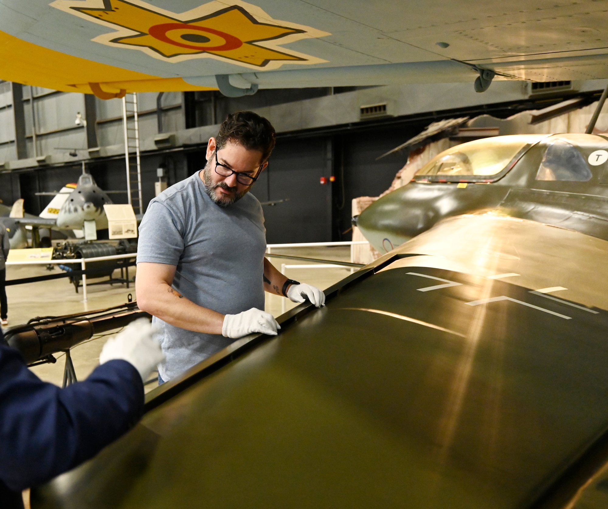 Man in a grey t-shirt standing next to air aircraft wing wearing white gloves, runs his hands down a green aircraft wing to feel the curve.