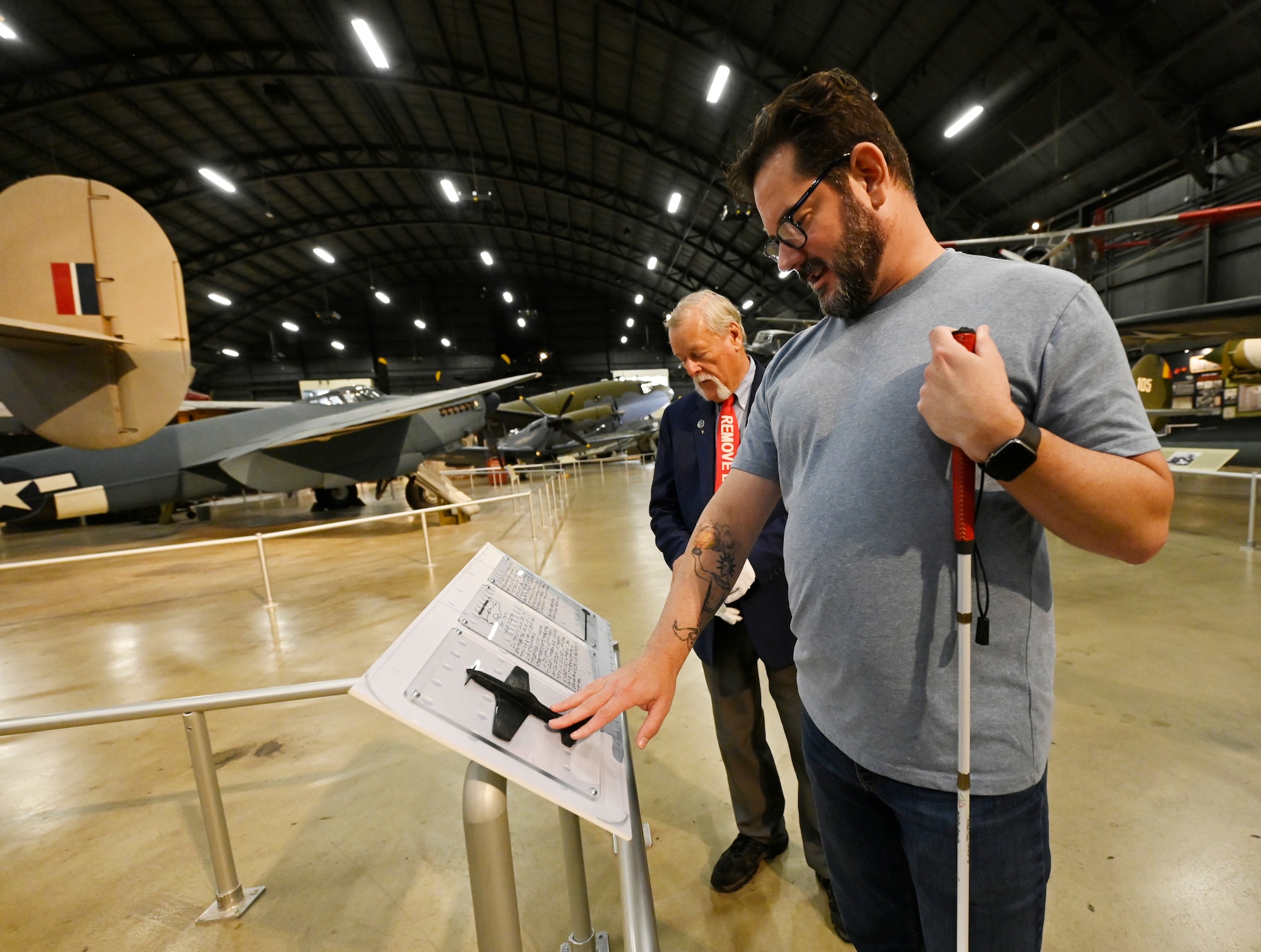 Man in grey t-shirt and jean holding a white cane in one hand and touching a 3-D model of an airplane attached to a sign that has the airplane's details printed in braille. A museum volunteer conducting the tour stands beside him.