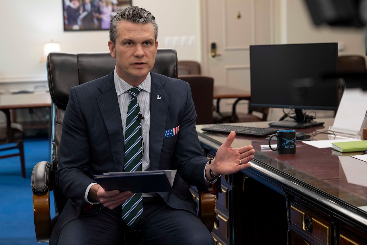 Defense Secretary Pete Hegseth delivers remarks from a chair while holding a notebook by a desk.