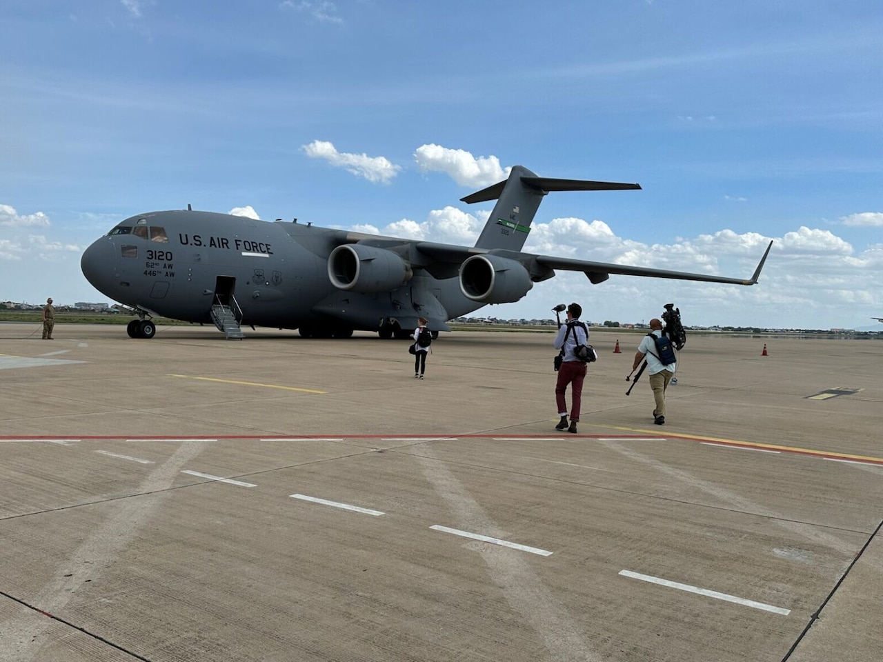 A large military airplane is on a tarmac while three people carrying camera equipment walk toward it.