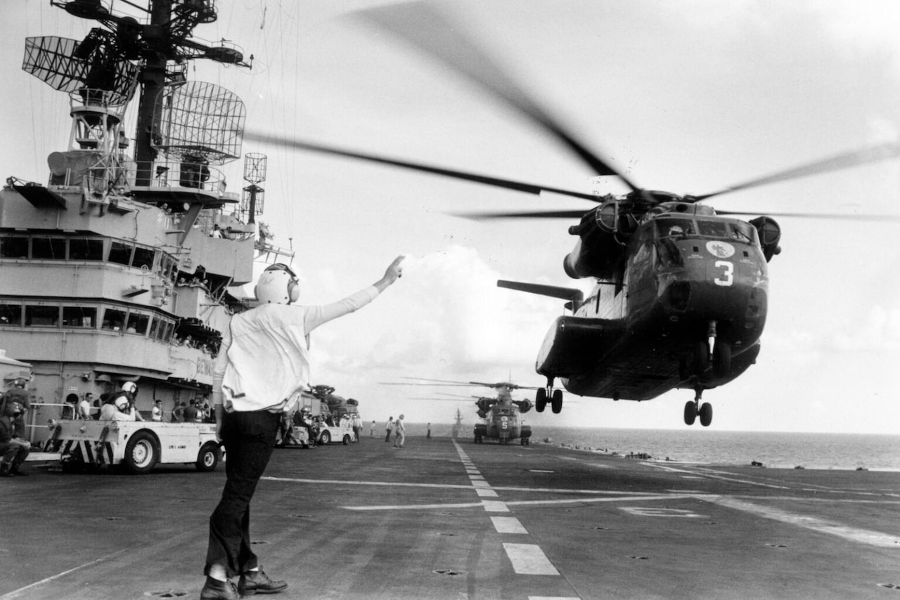 A man in uniform signals as a helicopter as it takes off from a naval vessel.
