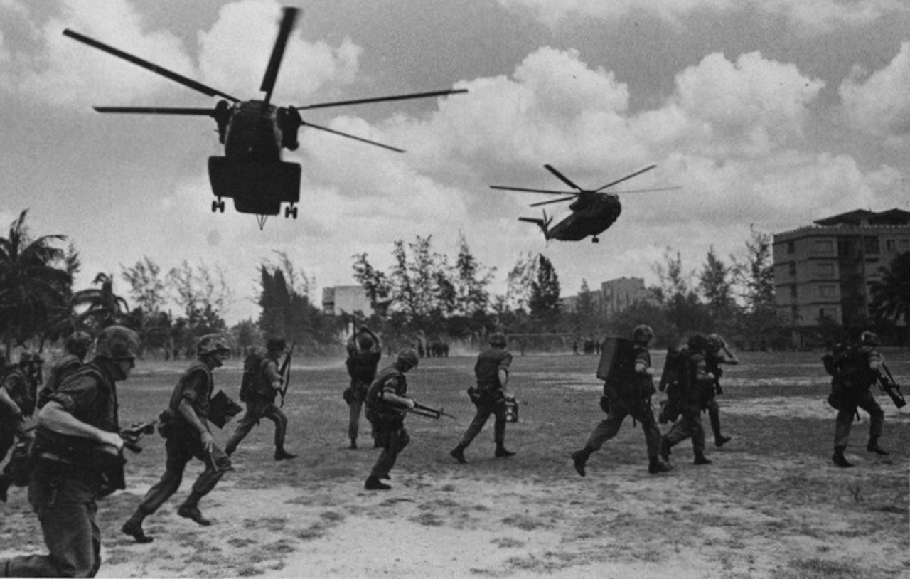 A dozen military personnel carrying weapons move across a field with two helicopters flying overhead.