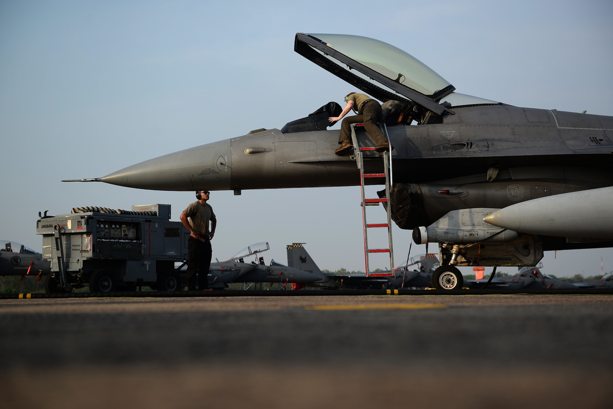 a mechanic leans into an open fighter jet cockpit while standing on a ladder