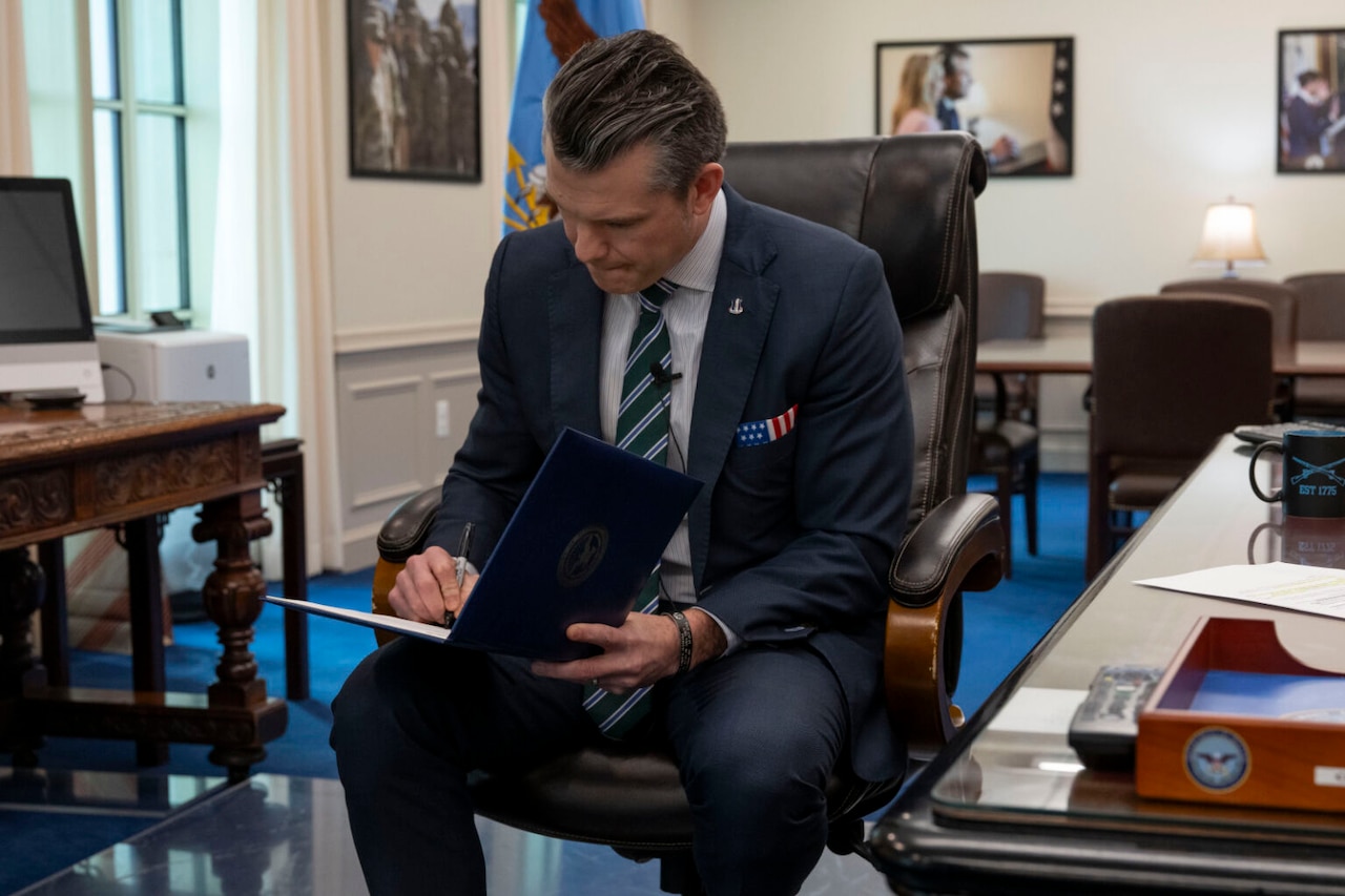 A man in a business suit is sitting in a chair in an office. He is holding a folder on his lap while signing a document contained inside it.