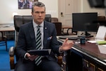 Defense Secretary Pete Hegseth delivers remarks from a chair while holding a notebook by a desk.