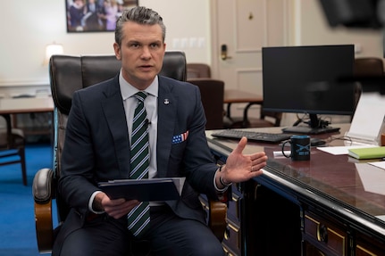 Defense Secretary Pete Hegseth delivers remarks from a chair while holding a notebook by a desk.