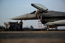 a mechanic leans into an open fighter jet cockpit while standing on a ladder