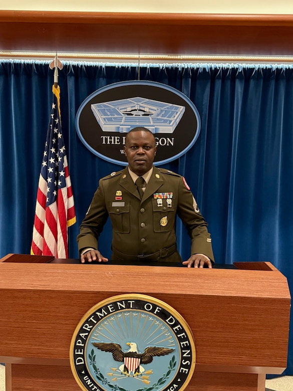 Male U.S. Army Soldier in dress uniform poses behind a Department of Defense podium with the Pentagon logo behind him