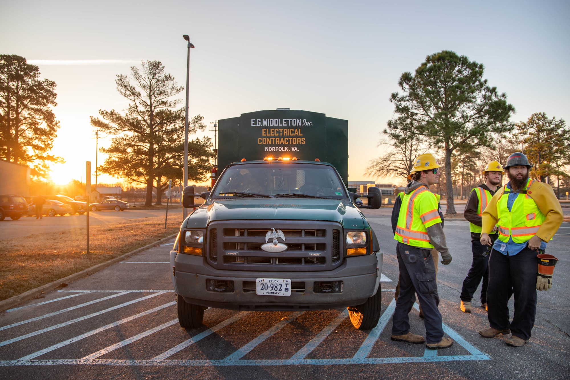 Men in hard hats and protective equipment stand next to work truck as the sun comes up in the background.