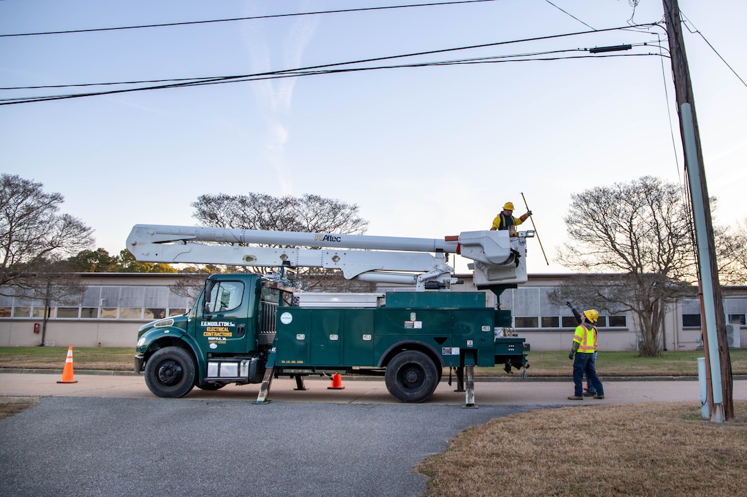 A lineman in a work truck hands a hot stick down to a coworker on the ground reaching up for it.