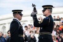 Two U.S. Army Soldiers in dark blue ceremonial dress uniforms face each other at the Tomb of the Unknown Soldier during the changing of the guard ceremony. One Soldier is inspecting a rifle while holding it horizontally in front of his face with both hands.