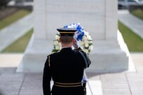 An Army Soldier dressed in dark blue ceremonial dress uniform is facing away from the camera while saluting towards a wreath that is place at the Tomb of the Unknown Soldier.