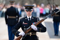 An Army Soldier in dark blue ceremonial uniform and wearing sunglasses is marching with a brown rifle in her hands at a 45-degree angle near the Tomb of the Unknown Soldier. There are two other Soldiers in the background standing facing the opposite direction.