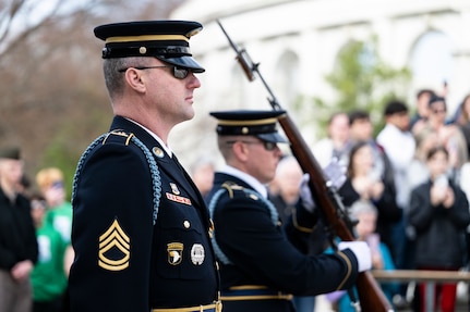 Two U.S. Army Soldiers in dark blue ceremonial dress uniforms are standing side by side with each other at the Tomb of the Unknown Soldier during the changing of the guard ceremony. One Soldier is adjusting his rifle while holding it vertically in front of his chest with both hands.