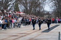 A large crowd (background) is watching three US Army Soldiers in dark blue ceremonial dress uniforms perform a changing of the guard ceremony at the Tomb of the Unknown Soldier.