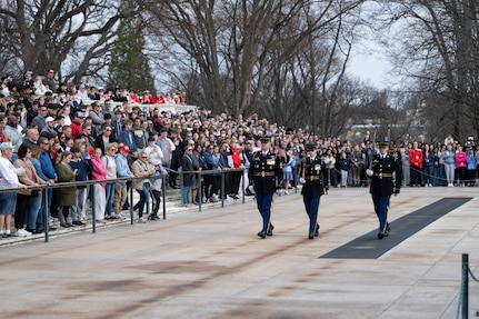 A large crowd (background) is watching three US Army Soldiers in dark blue ceremonial dress uniforms perform a changing of the guard ceremony at the Tomb of the Unknown Soldier.