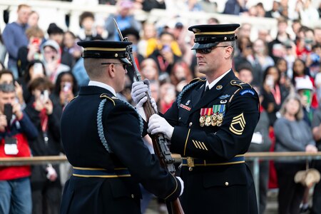Two U.S. Army Soldiers in dark blue ceremonial dress uniforms face each other at the Tomb of the Unknown Soldier during the changing of the guard ceremony. One Soldier is inspecting a rifle while handing it to the other Soldier with both hands and there is a large crowd watching in the background.