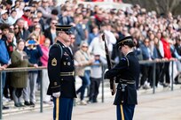 Two U.S. Army Soldiers in dark blue ceremonial dress uniforms face each other at the Tomb of the Unknown Soldier during the changing of the guard ceremony. One Soldier is inspecting a rifle while holding it vertically in front of her chest with both hands and there is a large crowd in the background.