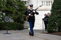 U.S. Army Soldier in dark blue ceremonial dress uniform is carrying a brown rifle in front of his chest as he marches down a stone walkway with large green bushes on the sides, towards the Tomb of the Unknown Soldier during the changing of the guard ceremony.