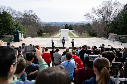 A large crowd (foreground) is watching three US Army Soldiers in dark blue ceremonial dress uniforms perform a changing of the guard ceremony at the Tomb of the Unknown Soldier.