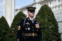 U.S. Army Soldier in dark blue ceremonial dress uniform is standing near the Tomb of the Unknown Soldier during the changing of the guard ceremony with tall green bushes in the background.