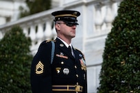 U.S. Army Soldier in dark blue ceremonial dress uniform is standing near the Tomb of the Unknown Soldier during the changing of the guard ceremony with tall green bushes in the background.