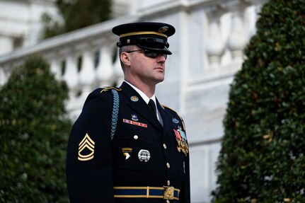 U.S. Army Soldier in dark blue ceremonial dress uniform is standing near the Tomb of the Unknown Soldier during the changing of the guard ceremony with tall green bushes in the background.