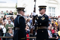 Two U.S. Army Soldiers in dark blue ceremonial dress uniforms face each other at the Tomb of the Unknown Soldier during the changing of the guard ceremony. One Soldier is inspecting a rifle while holding it vertically in front of his chest with both hands.