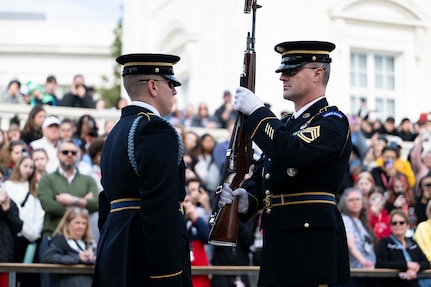 Two U.S. Army Soldiers in dark blue ceremonial dress uniforms face each other at the Tomb of the Unknown Soldier during the changing of the guard ceremony. One Soldier is inspecting a rifle while holding it vertically in front of his chest with both hands.