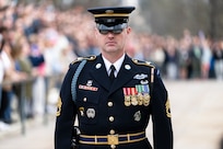 U.S. Army Soldier in dark blue ceremonial dress uniform is standing near the Tomb of the Unknown Soldier during the changing of the guard ceremony with dozens of onlookers in the distance.
