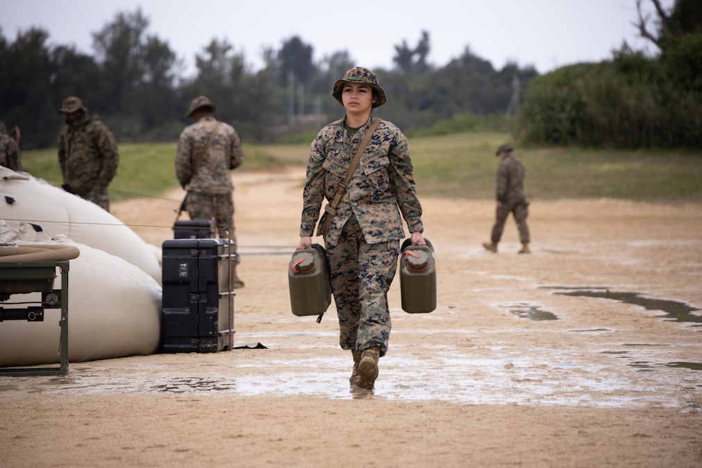 A Marine carrying two gas canisters while walking through mud as fellow service members work in the background.