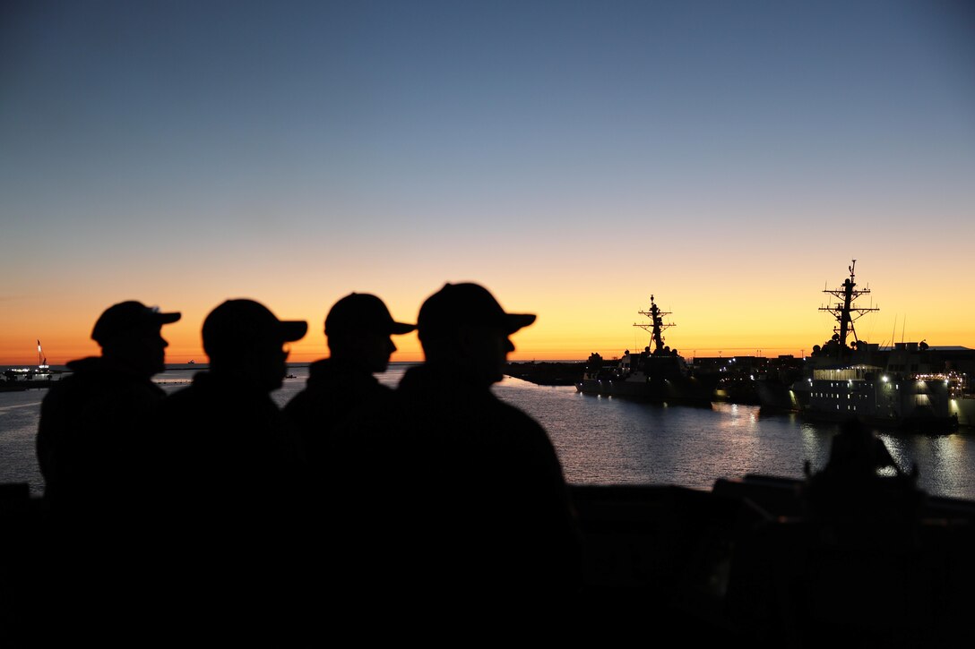 Four sailors look to the right while standing on a ship an seen in silhouette.