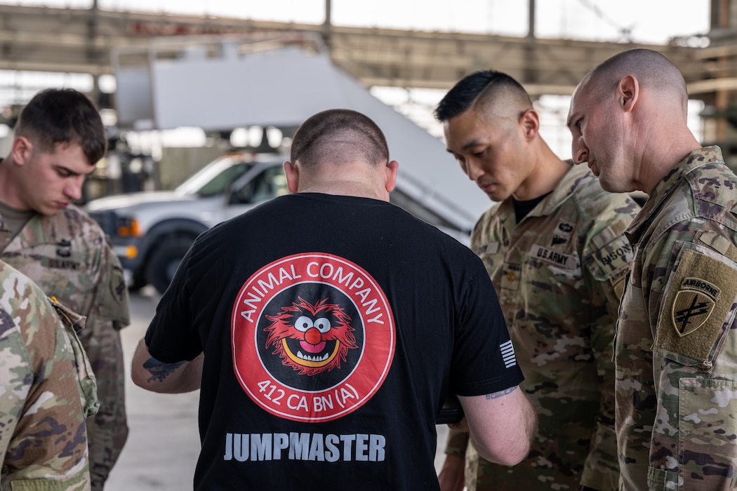 A member wearing a black t-shirt with the muppet Animal's face reading "Animal Company 412 CA BN (A) Jumpmaster" stands in the middle of three Army soldiers in military uniforms