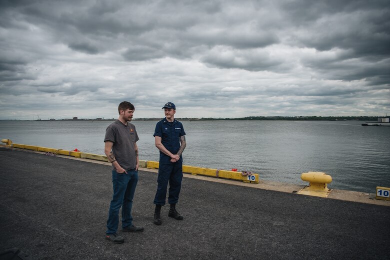Two men stand on a concrete boat dock. The individual on the left wears a gray t-shirt and stands with his hands in his pockets, while the individual on the right wears a navy blue U.S. Coast Guard uniform and hat.