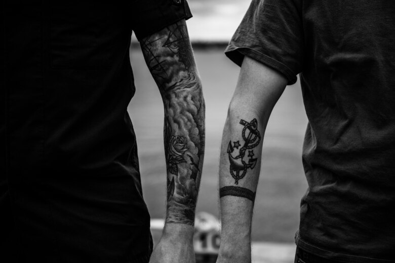 A black and white photo shows two individual's nautical tattoos. The left tattoo is a full sleeve of waves, while the right is an anchor with rope.